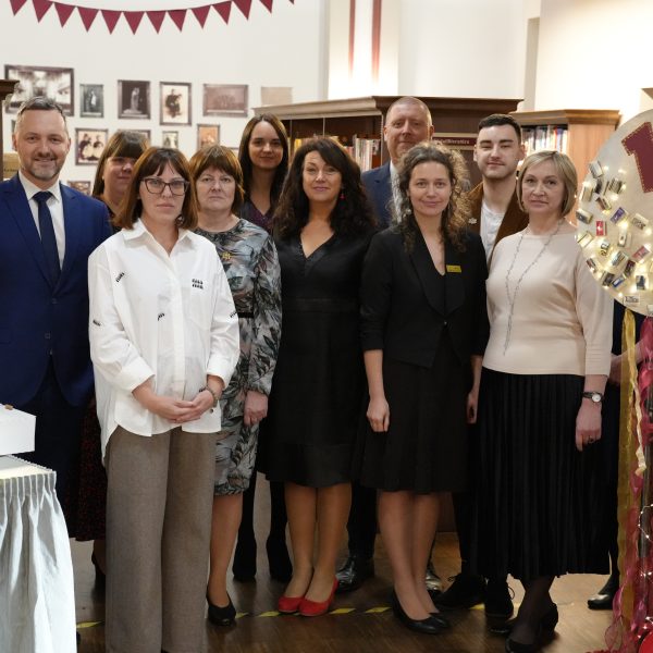 Vienpadsmit cilvēku grupa, trīs vīrieši un astoņas sievietes, pozē fotogrāfijai bibliotēkā. Kuldīgas Galvenās bibliotēkas kolektīvs 105.dzimšanas dienas svinībās.
