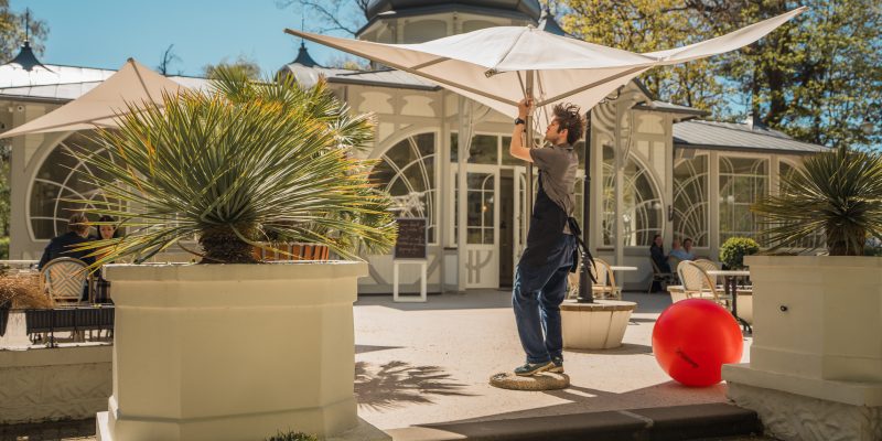 Vīrietis pelēkā kreklā un melnā priekšautā uzstāda lielu, gaišu saulessargu uz saulainas āra terases. Aiz viņa redzama eleganta ēka ar arkveida logiem un kupolu. Terasi rotā podos stādīti augi un liela sarkana piepūšamā bumba.