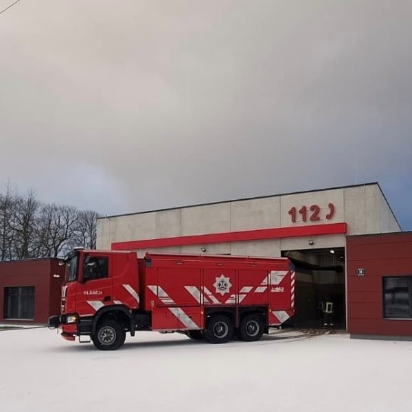 Sarkana ugunsdzēsēju mašīna ar baltām svītrām, uzrakstu "BLAŅES", emblēmu un numuru "402" novietota pie moderna ugunsdzēsēju depo ēkas, uz kuras fasādes redzami lieli sarkani cipari "112". Zeme ir apsnigusi.