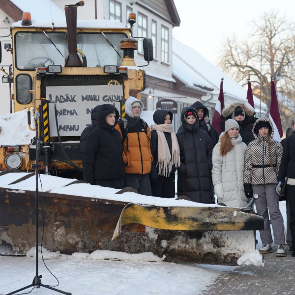 Grupa cilvēku, tostarp jaunieši, ziemas apģērbā stāv sniegotā laukumā pie liela dzeltena sniega tīrītāja ar uzgriezni priekšā. Daži no viņiem tur Latvijas karogus. Uz sniega tīrītāja ir zīme ar tekstu "LABĀK MANU GALVU ŅĒMA N MANU".