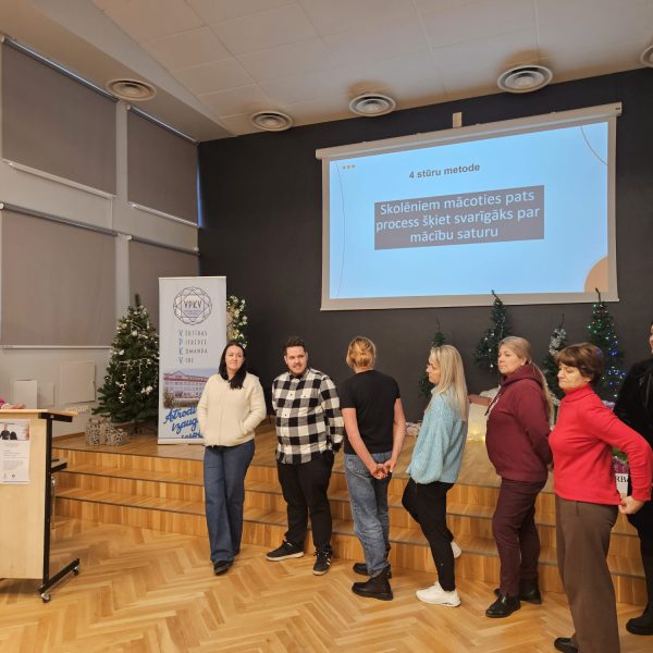 Sieviete rozā džemperī runā no pults auditorijas priekšā, kurā stāv vairākas sievietes un vīrieši.