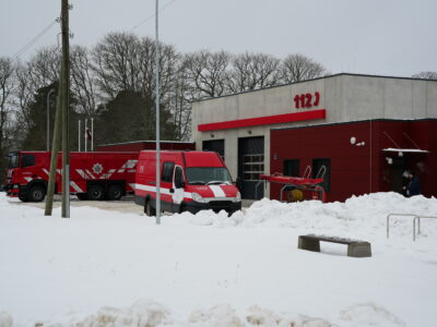 Sarkani-pelēka ugunsdzēsēju depo ēka ar uzrakstu "112" uz fasādes, ietverta sniegā. Pie tās stāv liela sarkana ugunsdzēsēju automašīna ar uzrakstu "OLAINES" un mazs sarkani balts mikroautobuss ar "112" un "2412" uz sāniem. Cilvēks redzams pie ēkas ieejas, fonā kaili koki.