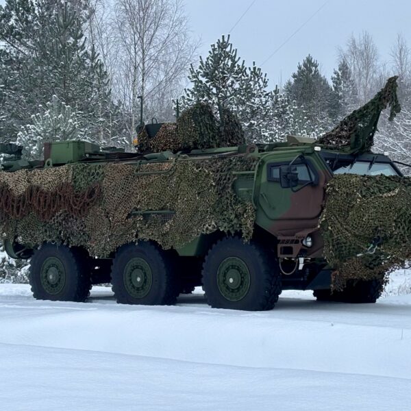 Bruņots zaļš militārais transportlīdzeklis ar maskēšanās tīklu stāv uz sniega klātas zemes ziemas mežā.
