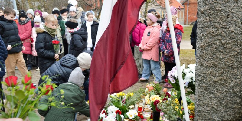 Bērni noliek ziedus un iedegtas sveces pie akmens pieminekļa, kura pakājē jau izvietoti daudzi ziedi un svecītes. Blakus plīvo Latvijas karogs. Fonā stāv vēl citi bērni un pieaugušie. Komunistiskā genocīda upuru piemiņas pasākumi novadā