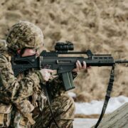 Notupies karavīrs kamuflāžas uniformā ar Latvijas karoga plāksteri un ķiveri mērķē ar šauteni ar optisko tēmekli, valkājot dzirdes aizsargus.