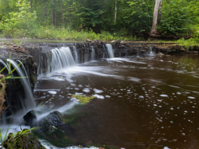 Ūdenskritums ar plūstošu, baltu ūdeni, kas krīt pāri tumšiem akmeņiem brūnganā upē, ko ieskauj zaļš mežs.